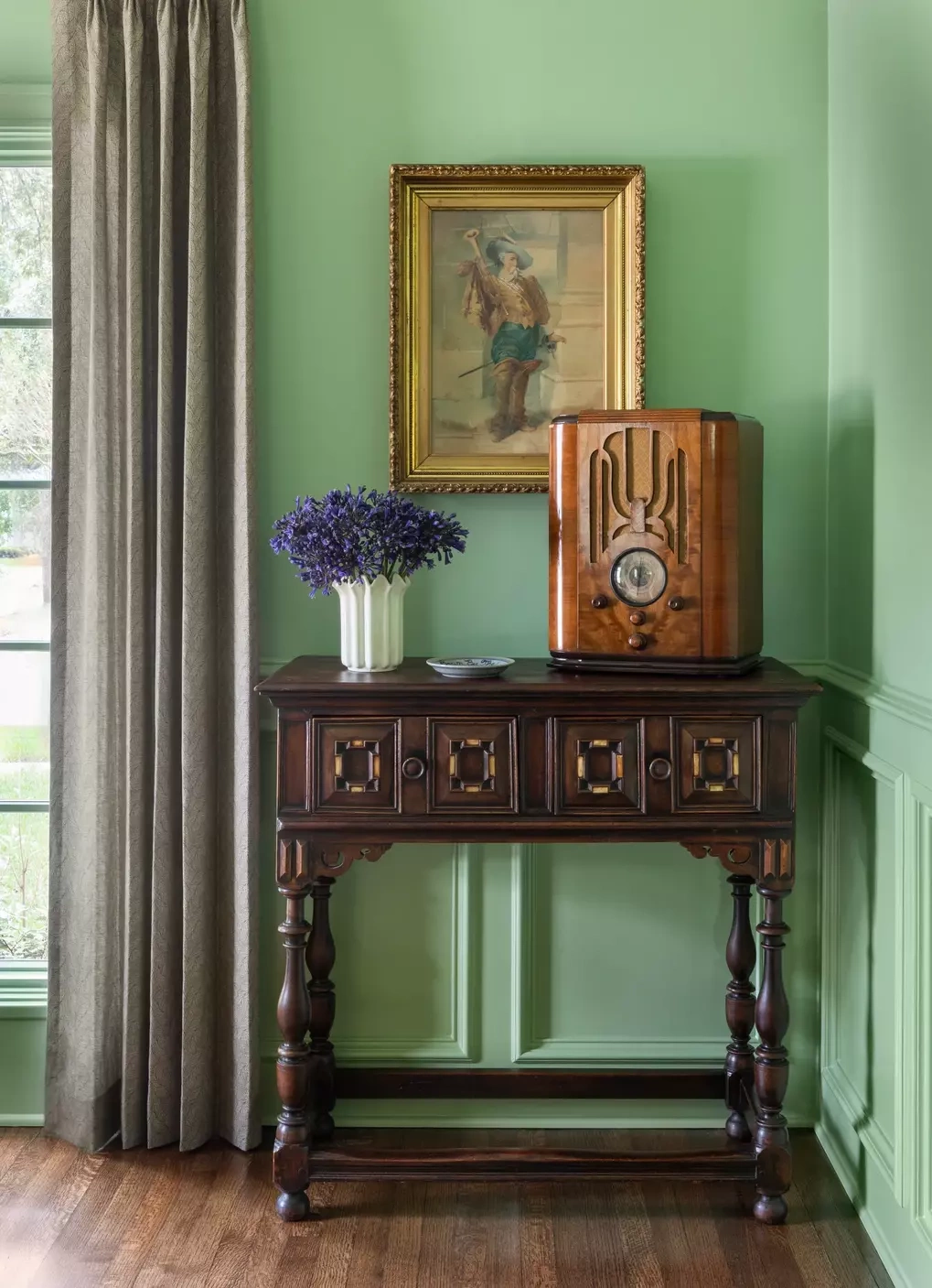 Chiếc radio cổ đặt trên bàn console gỗ trở thành điểm nhấn của góc phòng do Emily Spanos thiết kế. Tường xanh nhạt, rèm vải màu xám, tranh khung mạ và đồ gỗ tối màu tạo sắc thái hoài cổ, trong khi thiết bị nghe đài cơ học thay cho màn hình giúp không gian giữ tinh thần analog, giảm sự hiện diện của công nghệ số. Ảnh: Claudia Casbarian.